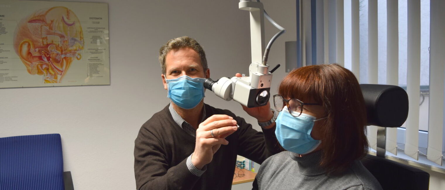 Ein Mann mit Mundschutz sitzt an einem Gerät zur Untersuchung des Gehörs einer Frau mit Mundschutz und Brille.