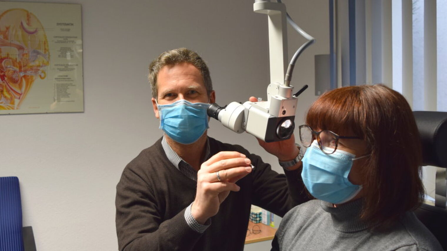 Ein Mann mit Mundschutz sitzt an einem Gerät zur Untersuchung des Gehörs einer Frau mit Mundschutz und Brille.