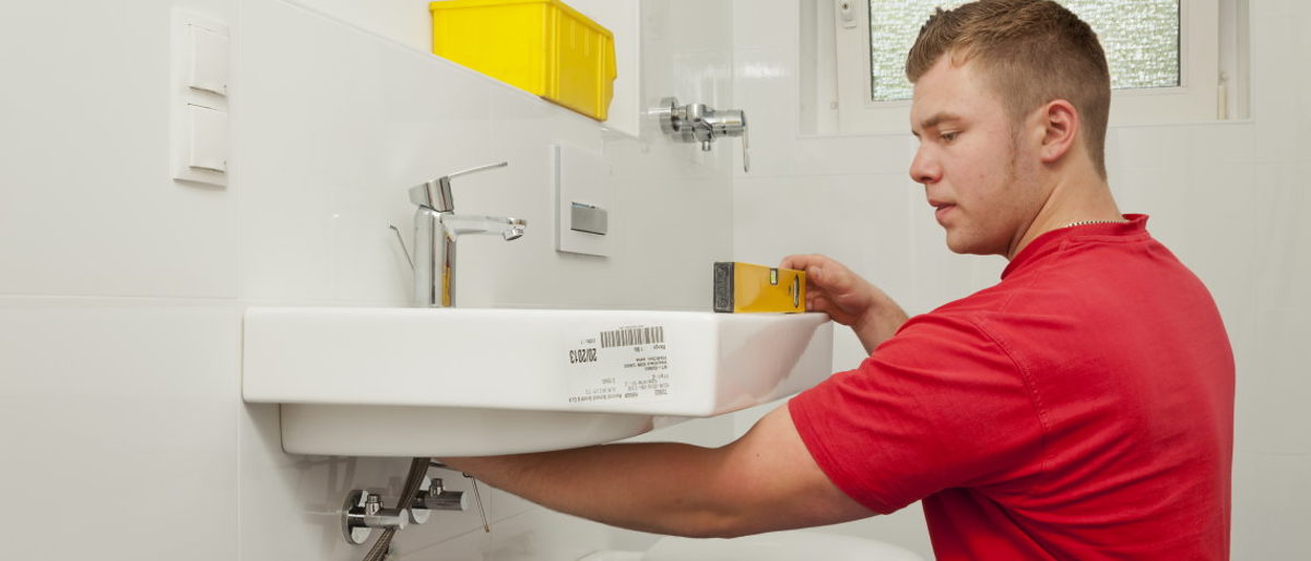 Barrierefreie Bäder erhöhen den Komfort in den eigenen vier Wänden. Ein junger Mann montiert ein Waschbecken in einem Badezimmer und überprüft mit einer Wasserwaage die korrekte Position.