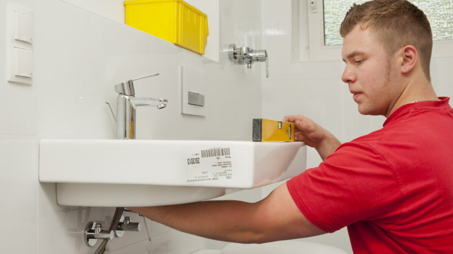 Ein junger Mann montiert ein Waschbecken in einem Badezimmer und überprüft mit einer Wasserwaage die korrekte Position.