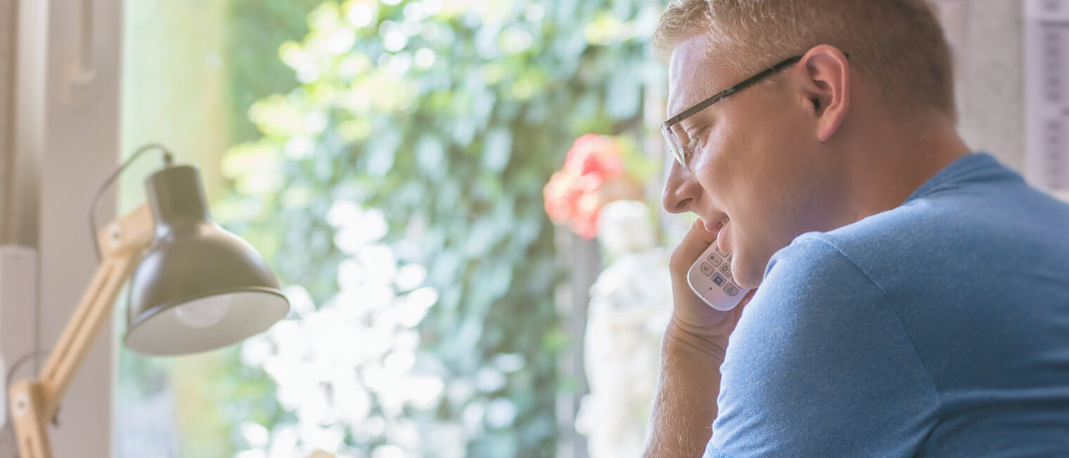 Eltern können sich über eine neue Hotline an die Berater wenden. Mann mit kurzen blonden Haaren und Brille telefoniert mit einem schnurlosen Telefon. Im Hintergrund Tischlampe und Fenster.