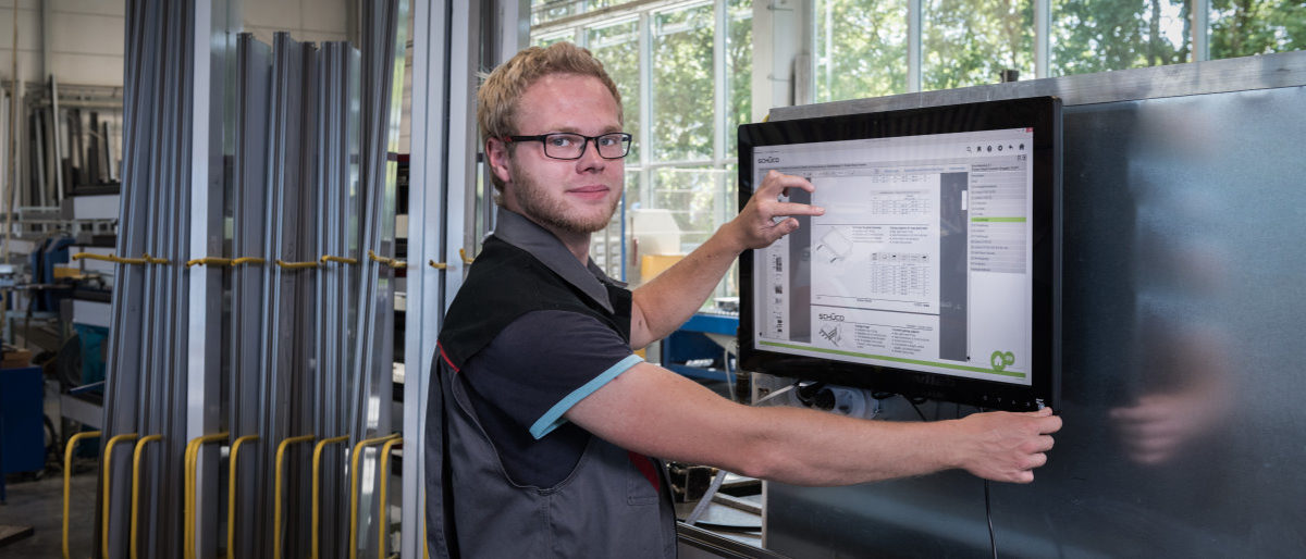 Innovationspreis BW - Aufmerksamkeit und Würdigung für Innovative Ideen Junger Handwerker in der Werkstatt arbeitet am Touch-Monitor und blickt in die Kamera.