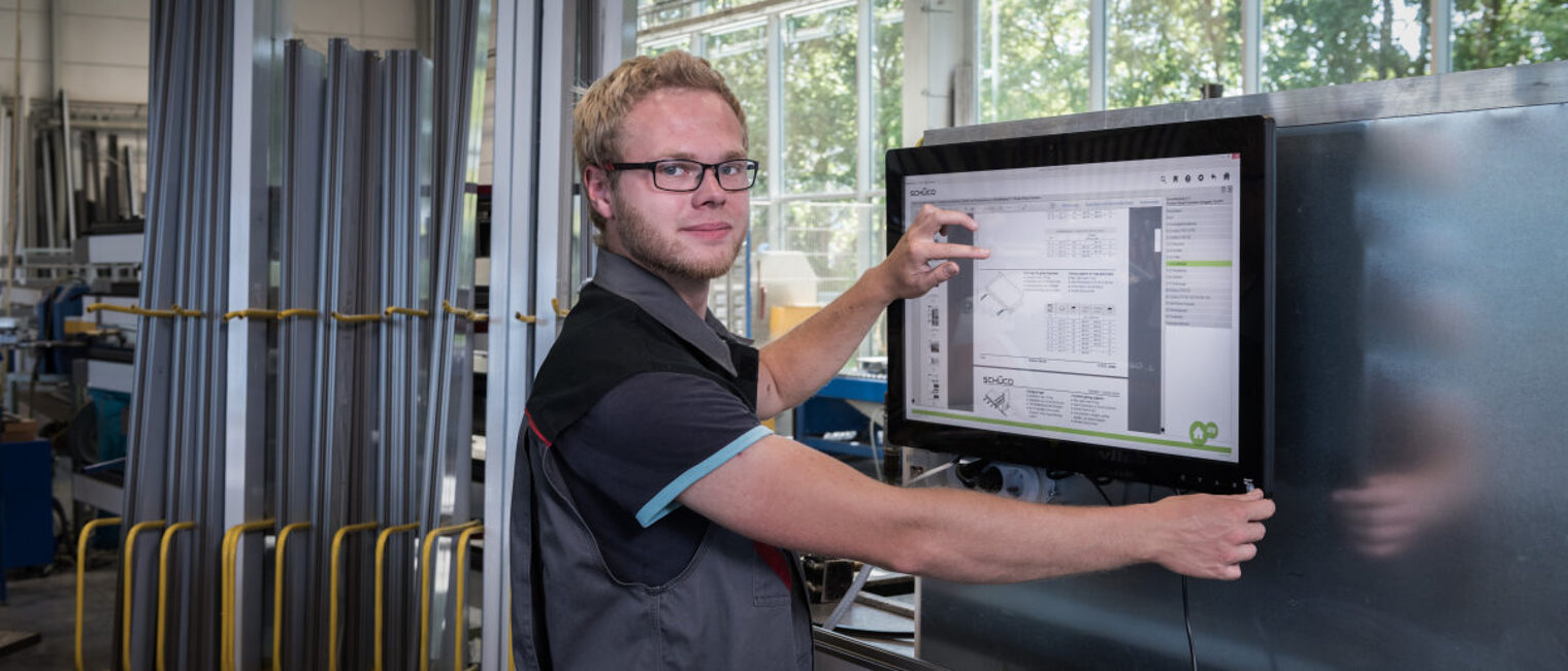 Junger Handwerker in der Werkstatt arbeitet am Touch-Monitor und blickt in die Kamera.