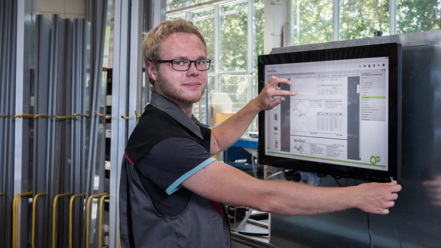 Junger Handwerker in der Werkstatt arbeitet am Touch-Monitor und blickt in die Kamera.