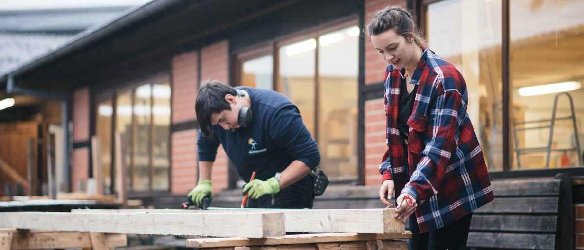 In Bauberufen können bei Praktika Abstände oft gut eingehalten werden. Junger Mann und junge Frau in Arbeitskleidung vermessen und markieren draußen vor einer Werkstatt einen Dachbalken.