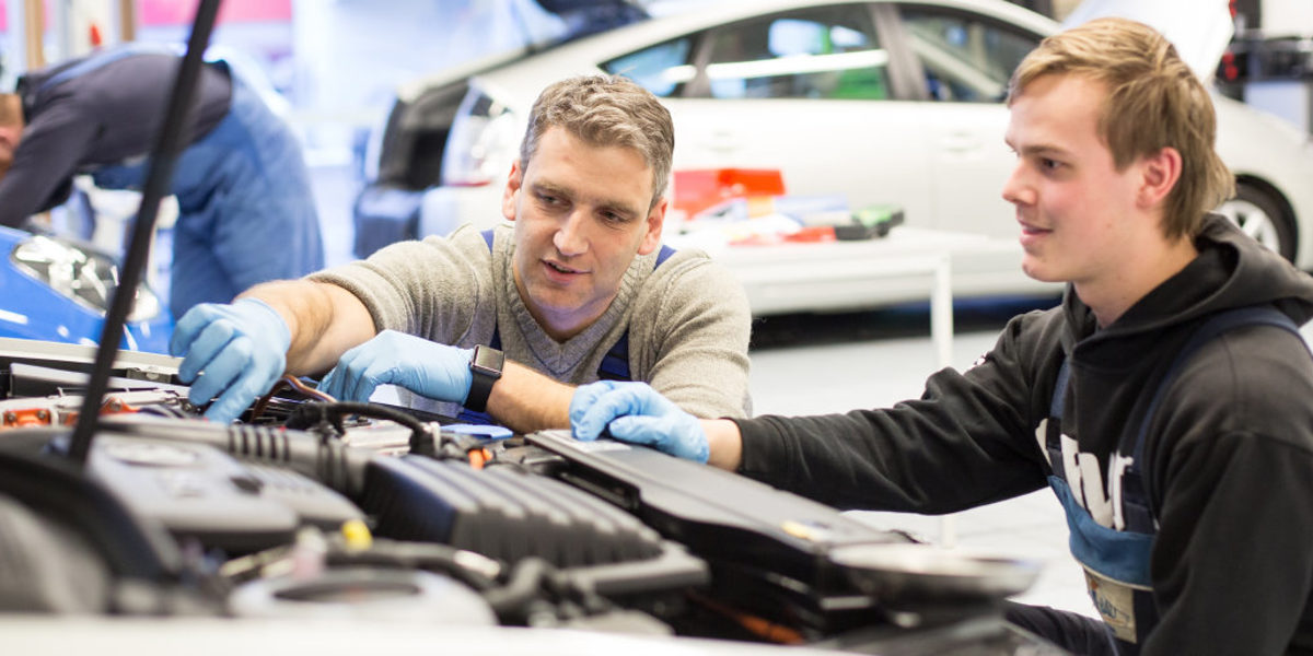 Im Ausbildertreff geht es um Führung und Motivation der Generation Z. Junger und älterer Mann in der Hocke vor dem Motorraum eines Autos in einer Werkstatt. Der ältere Mann zeigt und erklärt.