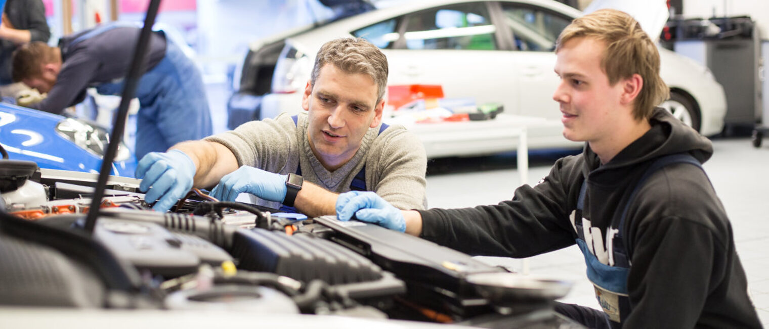 Im Ausbildertreff geht es um Führung und Motivation der Generation Z. Junger und älterer Mann in der Hocke vor dem Motorraum eines Autos in einer Werkstatt. Der ältere Mann zeigt und erklärt.