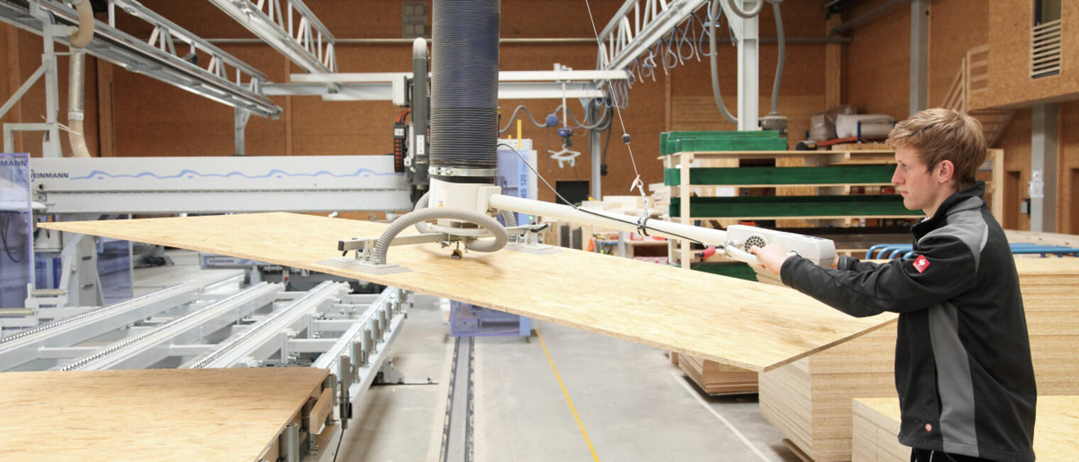 Trotz Corona laufen die Geschäfte bei der Mehrheit der Handwerker gut Junger Mann hebt mit Hilfe einer Maschine eine Platte aus Holz von einem Transportband in einer Fertigungshalle.