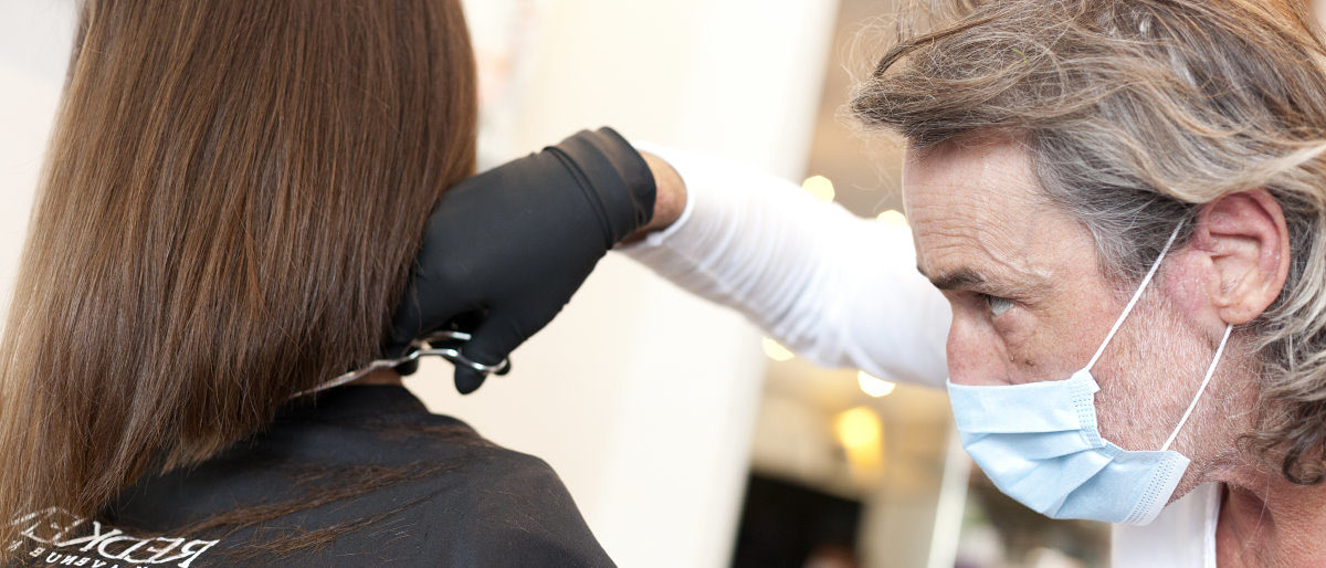 Neue Corona-Regeln für Friseurbetriebe und körpernahe Dienstleistungen Ein älterer Mann mit einem medizinischen Mund-Nasen-Schutz schneidet die Spitzen der langen braunen Haare einer Kundin.