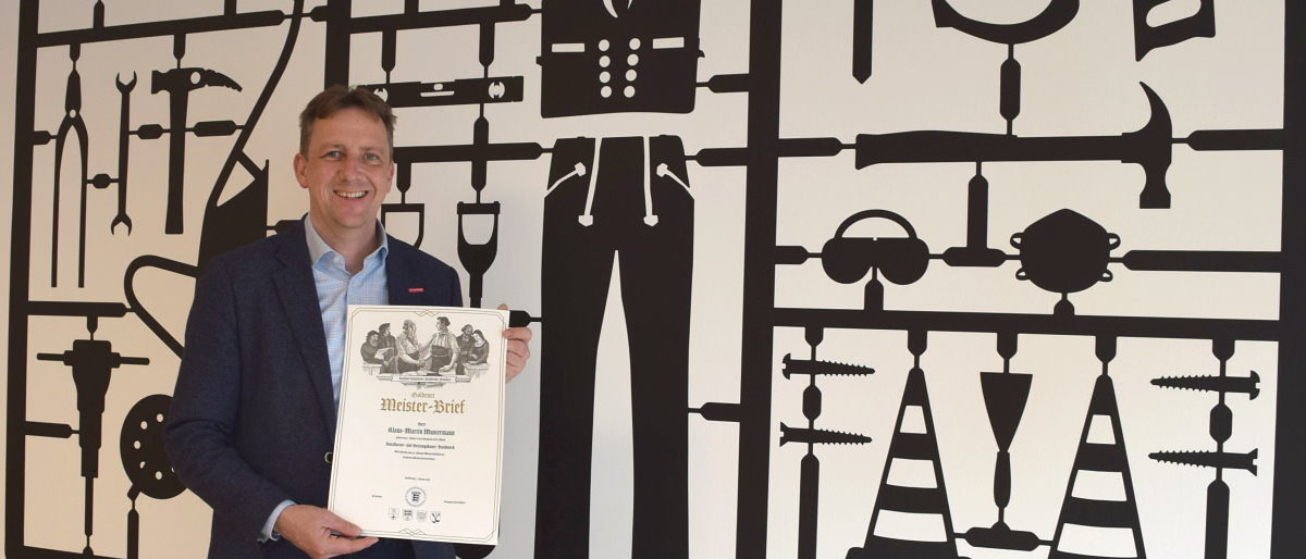 Hauptgeschäftsführer Ralf Schnörr präsentiert den Schmuckmeisterbrief. Mann in blauem Jackett steht vor einer Wand, auf der handwerkliche Gegenstände abgebildet sind und präsentiert ein Dokument.