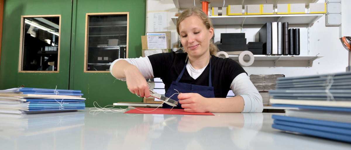 Das Buchbinderhandwerk ist immaterielles Kulturerbe in Deutschland. Junge Frau sitzt in Werkstatt an Tisch mit ungebundenen Bücherstapeln und arbeitet mit Schere und Faden an einem Buchrücken.