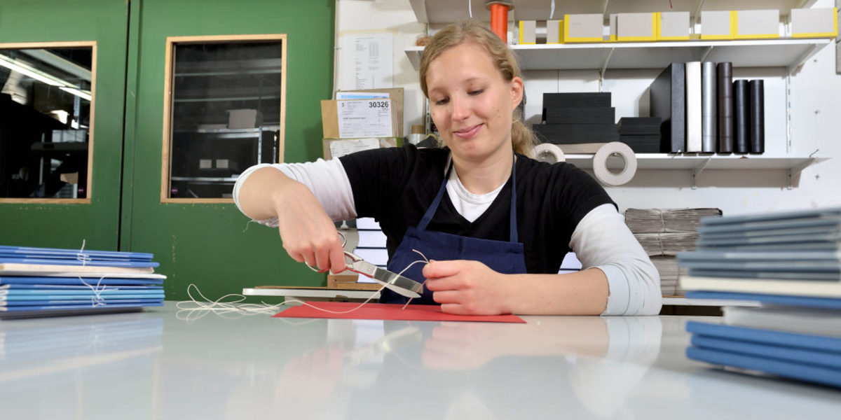 Das Buchbinderhandwerk ist immaterielles Kulturerbe in Deutschland. Junge Frau sitzt in Werkstatt an Tisch mit ungebundenen Bücherstapeln und arbeitet mit Schere und Faden an einem Buchrücken.