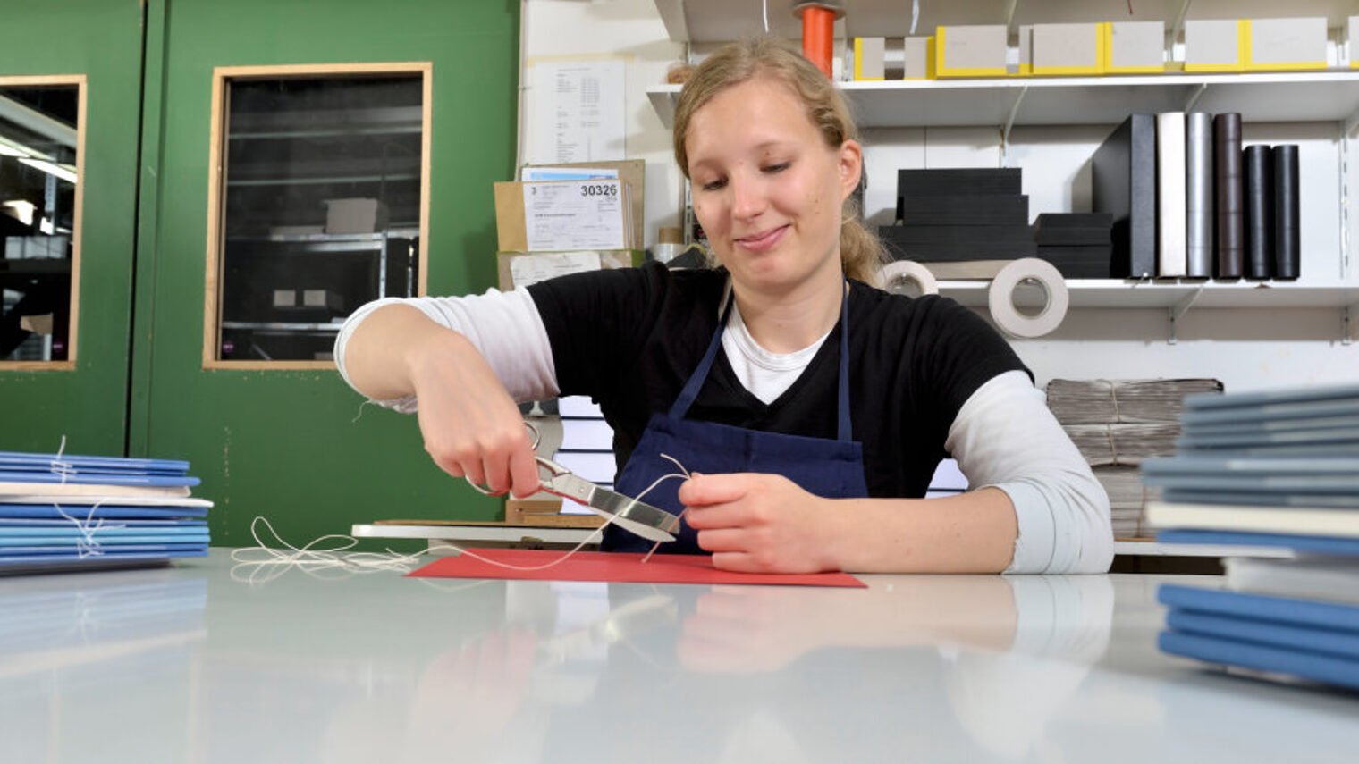 Junge Frau sitzt in Werkstatt an Tisch mit ungebundenen Bücherstapeln und arbeitet mit Schere und Faden an einem Buchrücken.