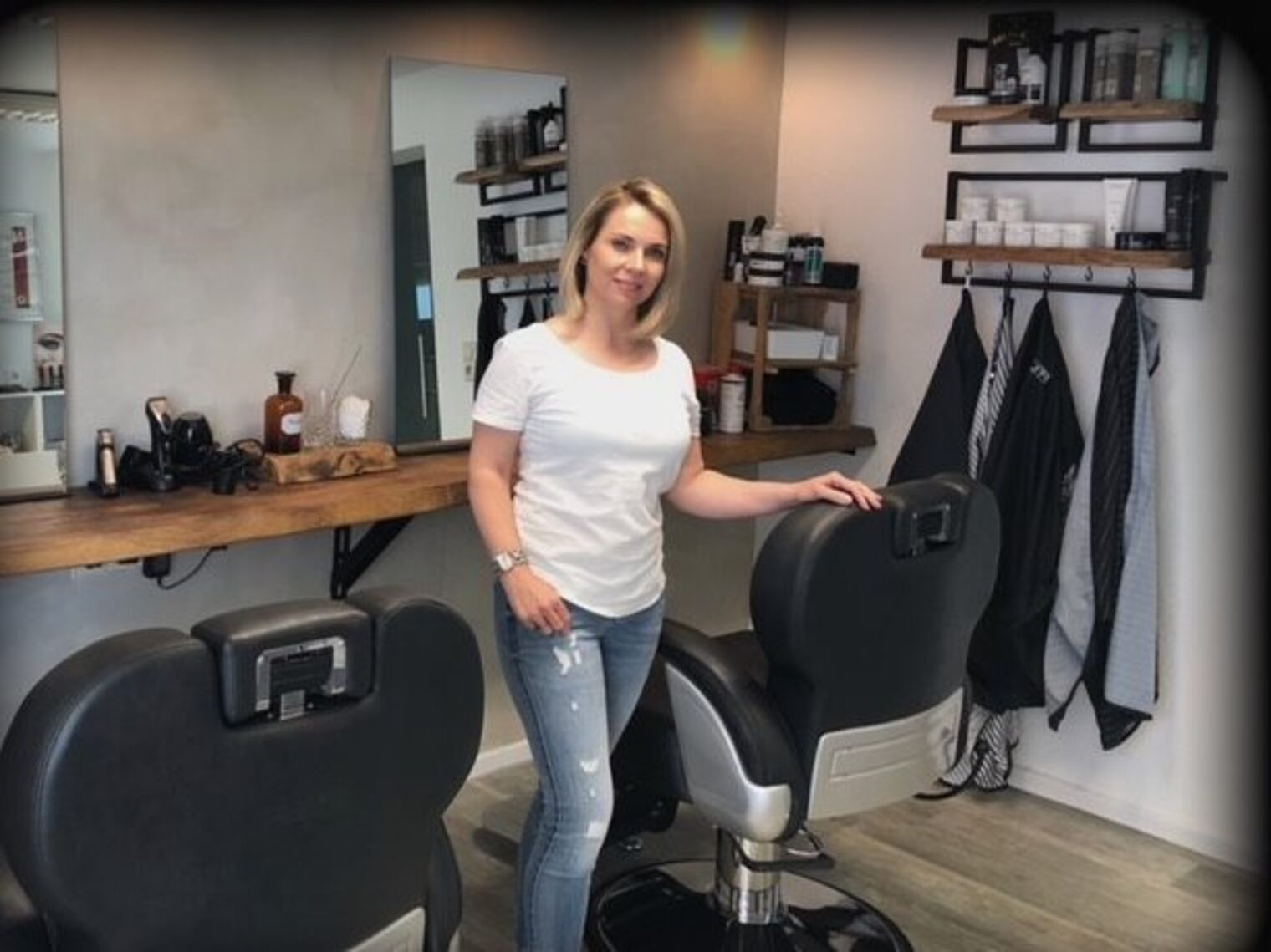 Frau mit blonden Haaren in Jeans und weißem T-Shirt zwischen zwei Stühlen in einem Friseursalon, blickt in die Kamera.