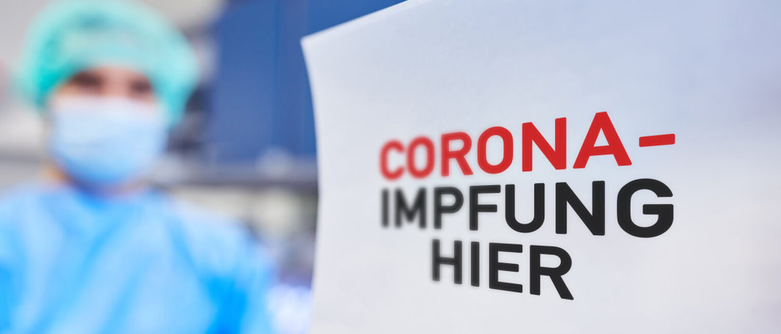 Rechts im Vordergrund ein weißes Schild mit Text "Corona-Impfung hier". Im Hintergrund unscharf eine Person Schutzkleidung