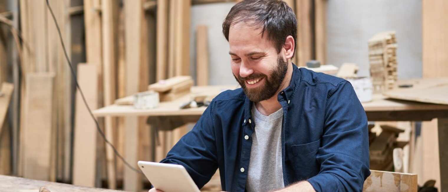 Mann sitzt in einer Schreinerwerkstatt und schaut lächelnd auf ein Tablet.