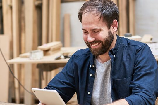 Digitalisierte Prozesse können helfen, die Effizienz zu steigern.  Mann sitzt in einer Schreinerwerkstatt und schaut lächelnd auf ein Tablet.