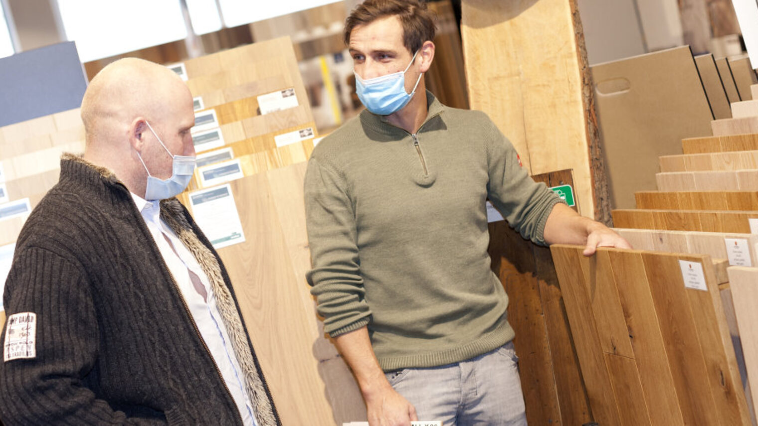Zwei Männer mit medizinischen Masken stehen in einem Raum mit vielen verschiedenen Ansichtsexemplaren von Parkettböden. 