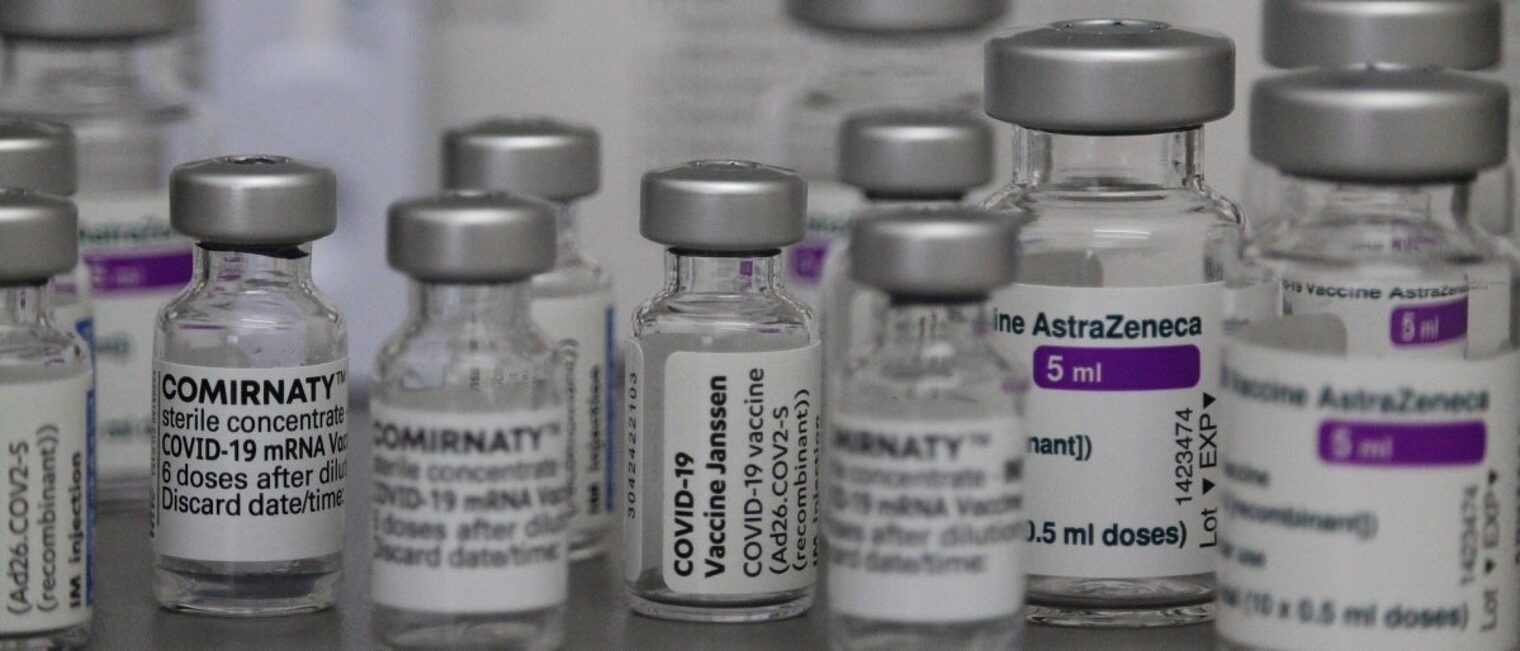 Kleine Fläschchen in verschiedenen Größen mit den Labels der Impfstoffe gegen Corona wie Comirnaty, Janssen und AstraZeneca.