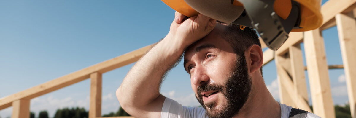 Ein Bauarbeiter steht schwitzend mit einem gelben Helm in der Hand auf einem Dachstuhl. 