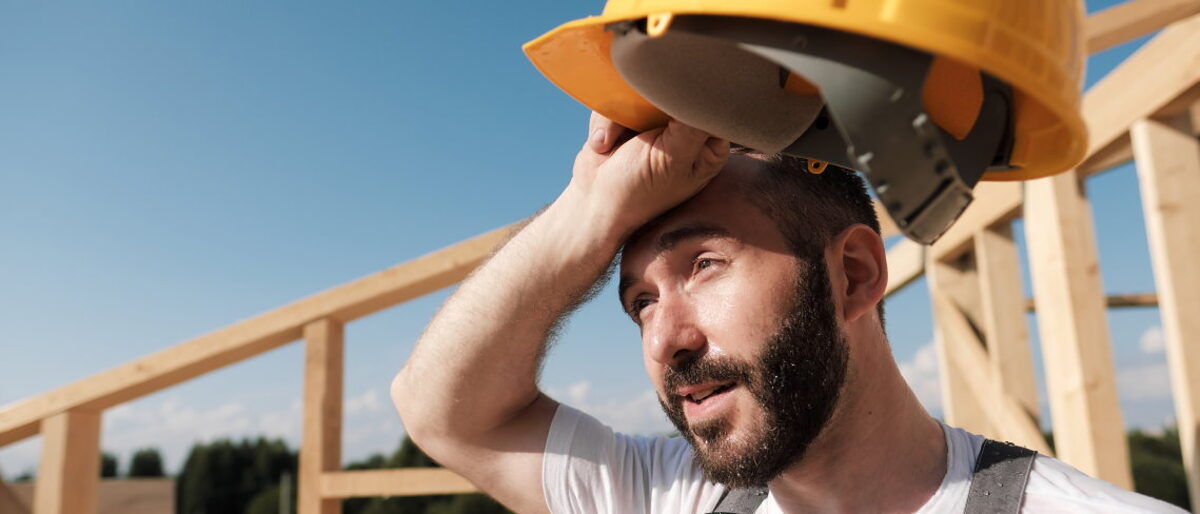 Ein Bauarbeiter steht schwitzend mit einem gelben Helm in der Hand auf einem Dachstuhl. 