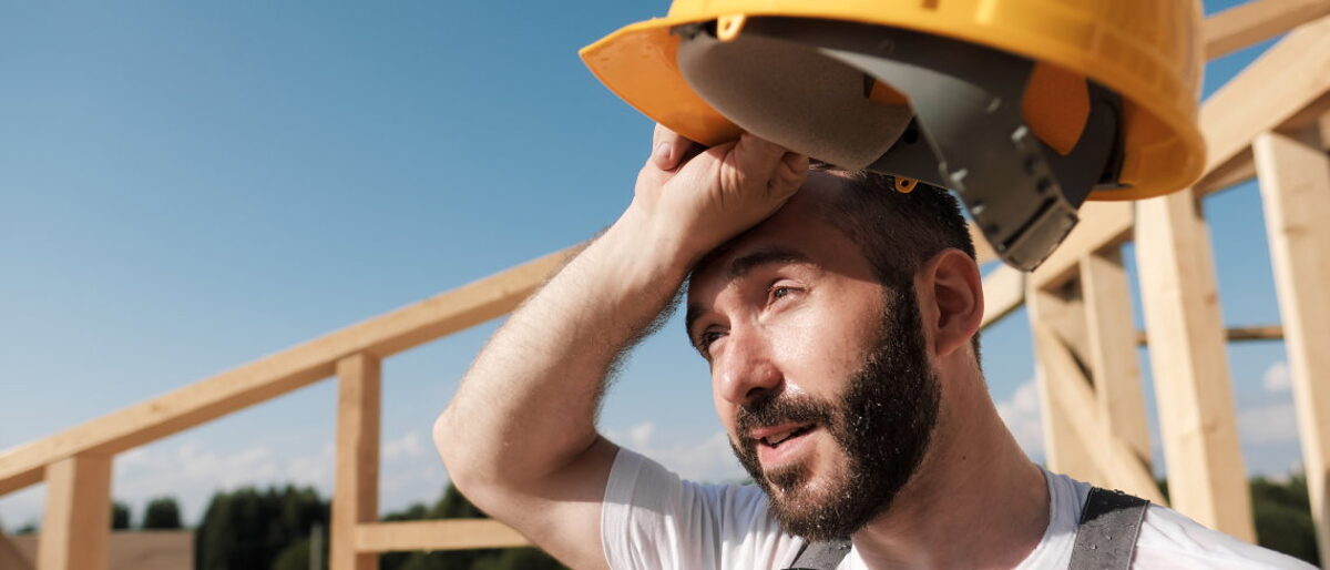 Der direkten Sonne ausgesetzt zu sein, ist eine Belastungsprobe für Hand-werker. Ein Bauarbeiter steht schwitzend mit einem gelben Helm in der Hand auf einem Dachstuhl.