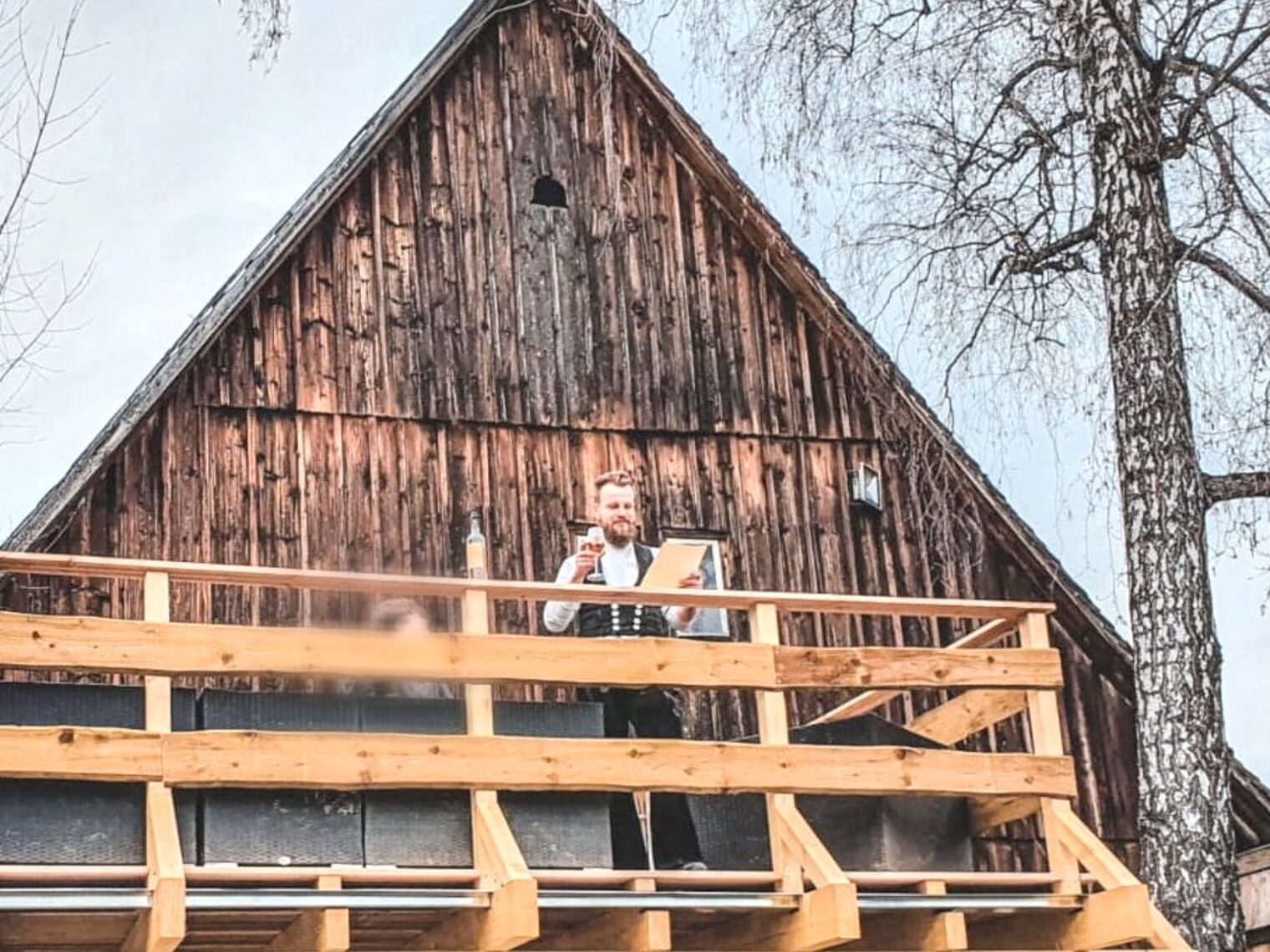 Ein Mann in Zimmererkluft steht auf einem Holzgerüst vor einer Hausfassade aus Holz mit Glas und Blatt Papier in der Hand. 