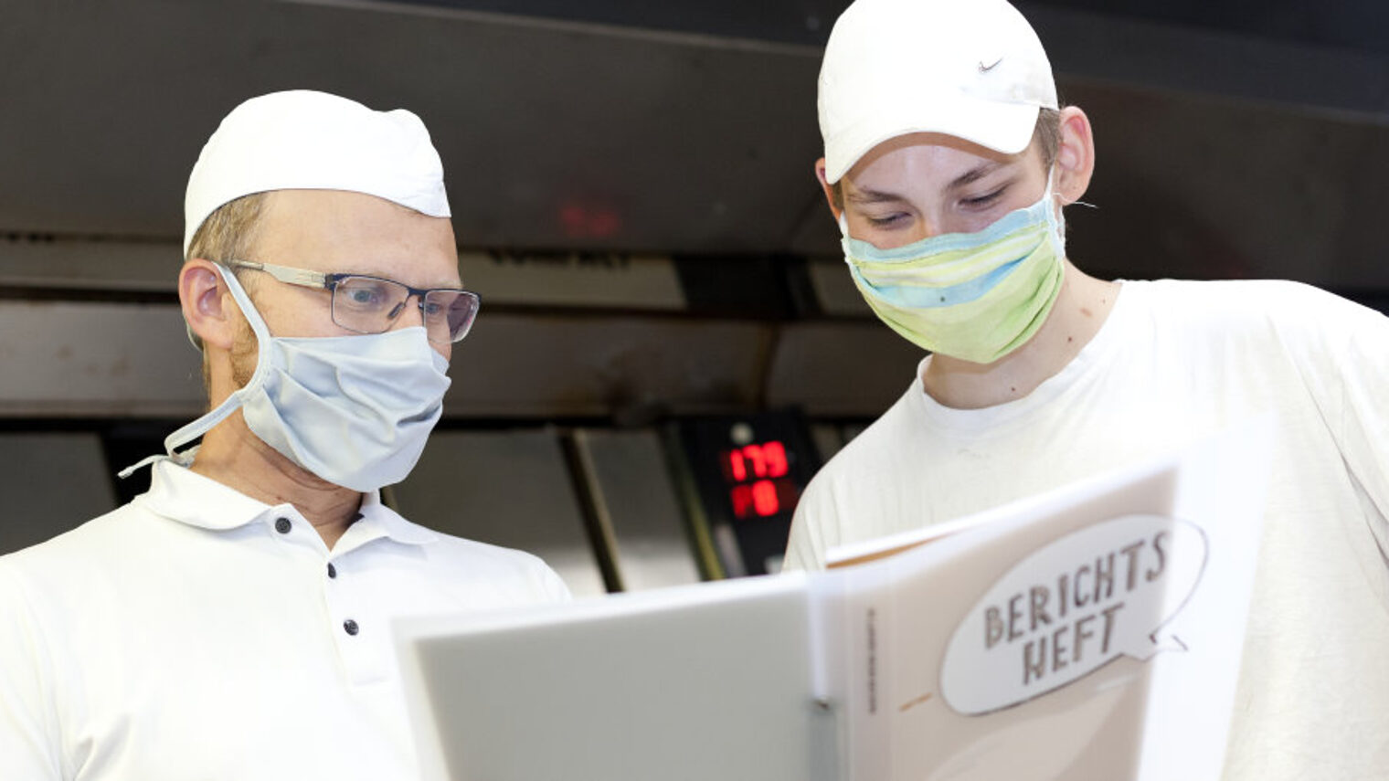 Zwei Männer in weißer Arbeitskleidung der Bäcker und Mund-Nasen-Schutz sehen in einen Ordner mit Aufschrift "Berichtsheft".