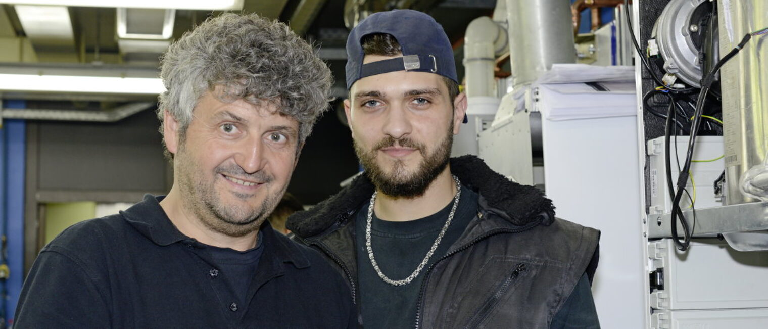 Zwei Fachkräfte, ein älterer und ein junger Handwerker bei der Arbeit schauen zufrieden in die Kamera.