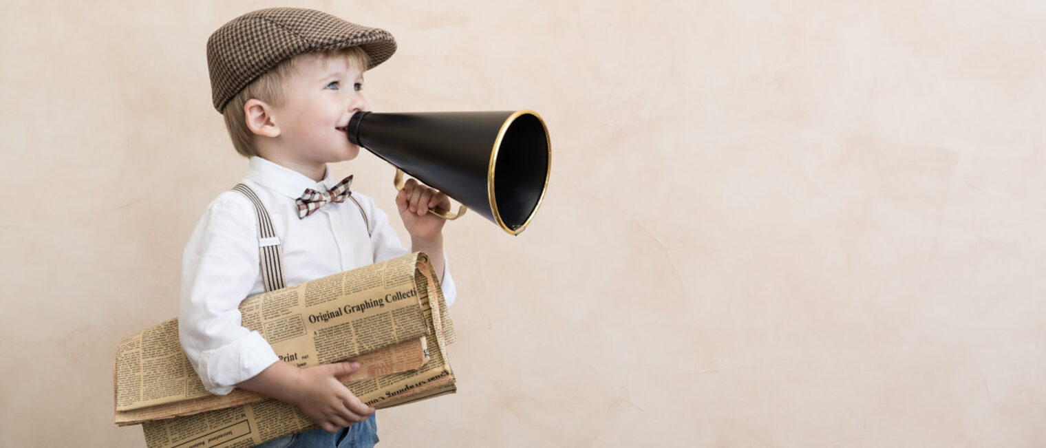 Kleiner Junge in altmodischer Kleidung hält eine Zeitung unter dem rechen Arm und ein Megafon in der linken Hand vor dem Mund