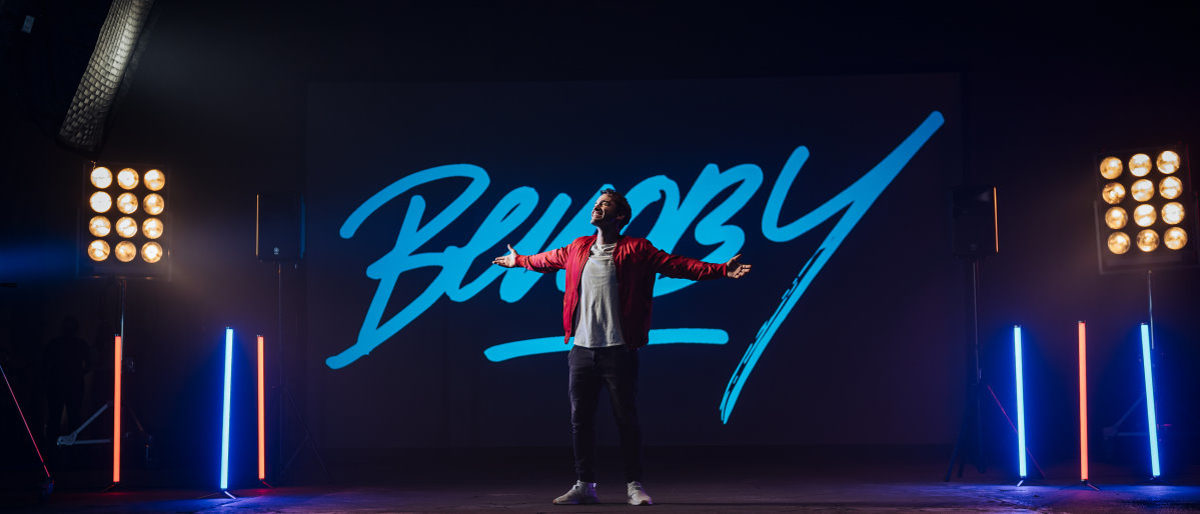 Benoby singt "Was für immer bleibt" - den neuen Song für das Handwerk. Ein Mann steht mit ausgebreiteten Armen auf einer Bühne, im Hintergrund Scheinwerfer, farbige Beleuchtung und ein Schriftzug.