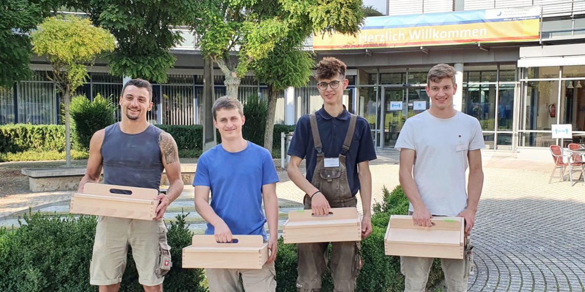 Kammersieger Felix Wilhelm (r.) mit seinen Tischler-Kollegen. Vier junge Männer stehen vor einem Gebäude im Freien. jeder hat einen Kasten aus Holz mit Griff oben in der Hand.