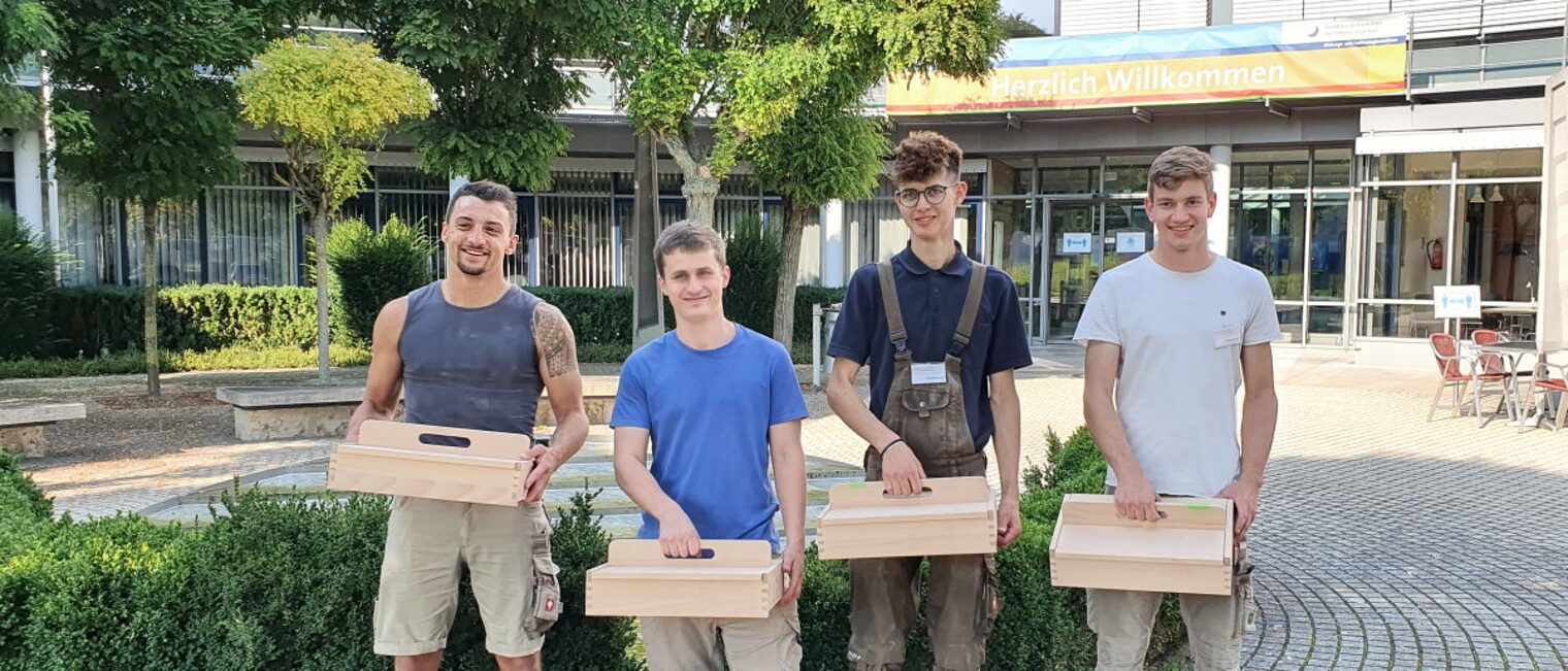 Vier junge Männer stehen vor einem Gebäude im Freien. jeder hat einen Kasten aus Holz mit Griff oben in der Hand.