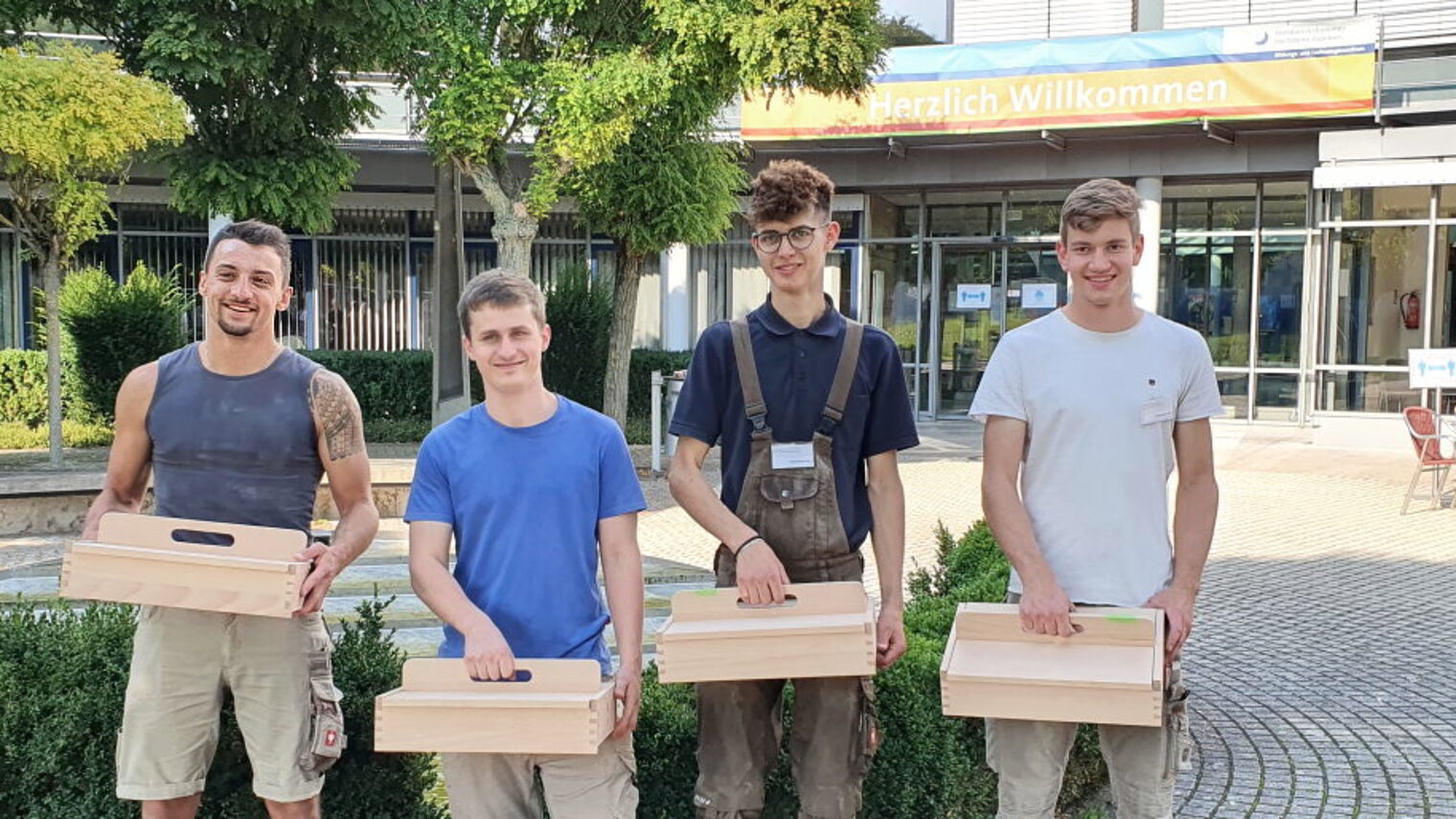 Vier junge Männer stehen vor einem Gebäude im Freien. jeder hat einen Kasten aus Holz mit Griff oben in der Hand.