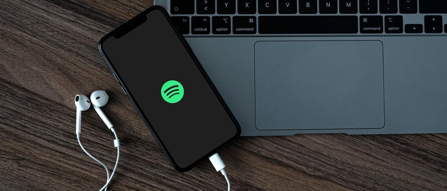 Neue Podcast-Reihe zum Thema internationale Fachkräfte finden. Auf einer Holzfläche steht ein Laptop, darauf ein Smartphone mit rundem grünen Symbol im Display und weißen In-Ear-Kopfhörern