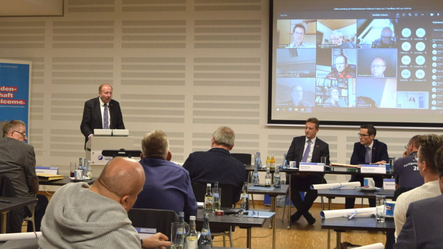 Mann steht an Rednerpult, mehrere Menschen sitzen an Tischen davor, daneben und auf einer Leinwand über Videokonferenz.