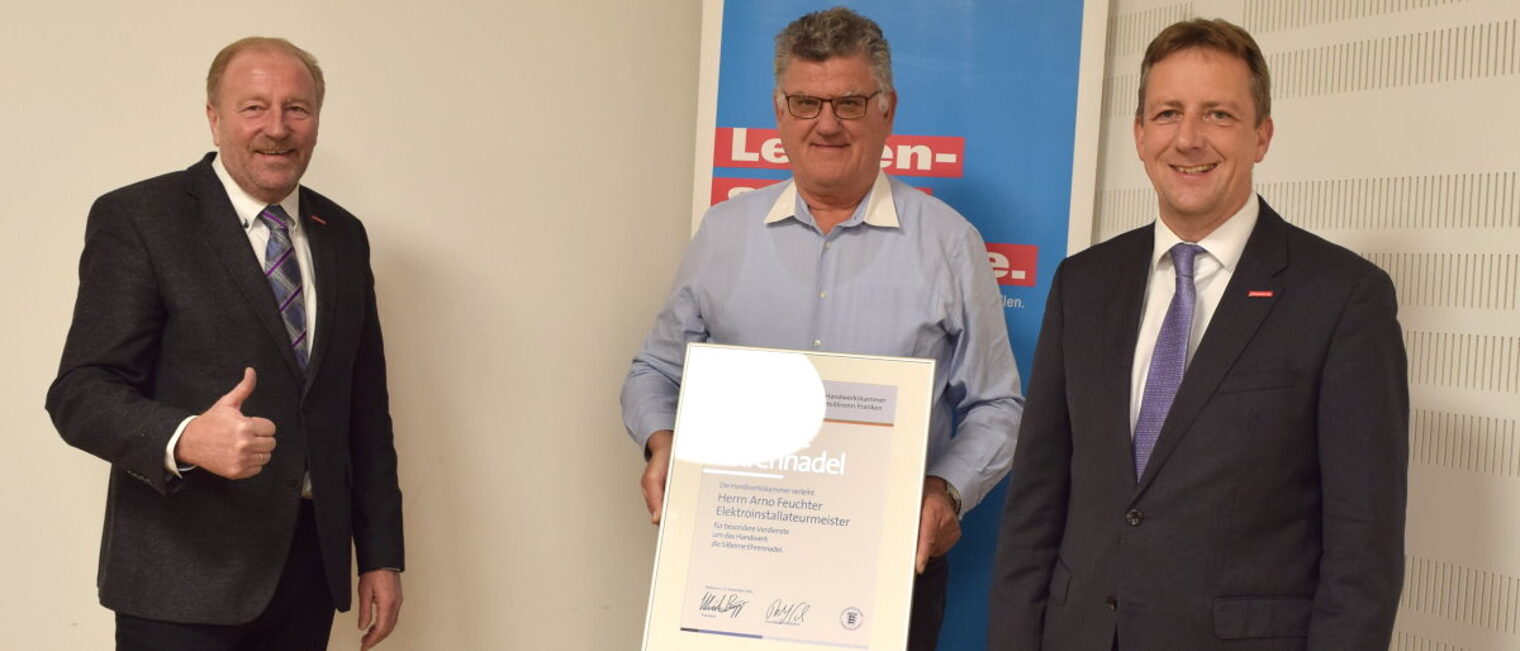 Arno Feuchter (M.) erhielt die silberne Ehrennadel der Handwerkskammer. Zwei Männer in Anzügen stehen neben einem Mann in ihrer Mitte, der eine gerahmte Urkunde in den Händen vor sich hält.