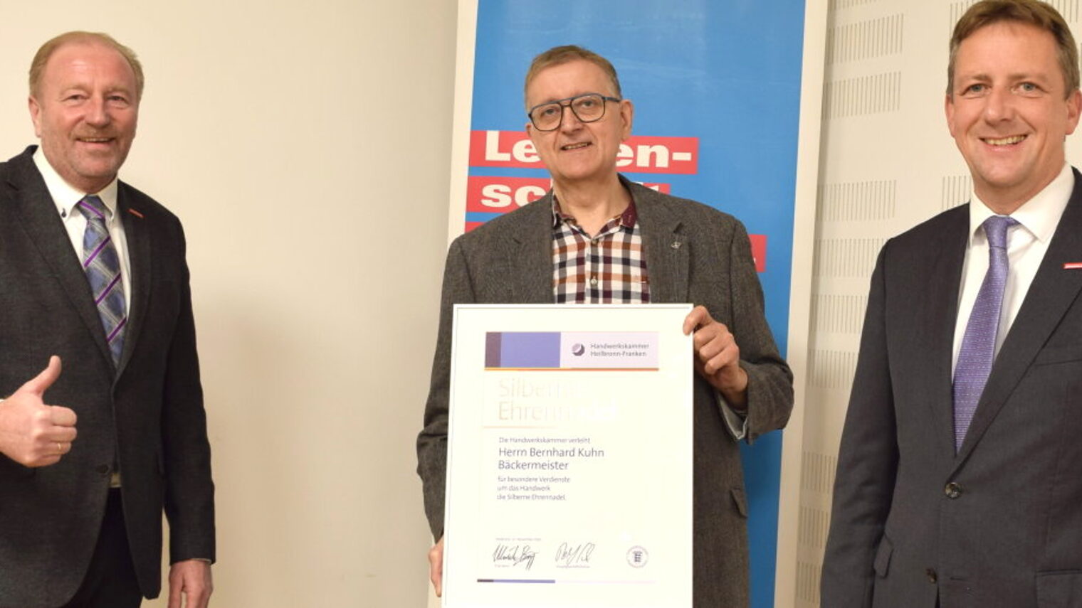Zwei Männer in Anzügen stehen neben einem Mann in ihrer Mitte, der eine gerahmte Urkunde in den Händen vor sich hält.