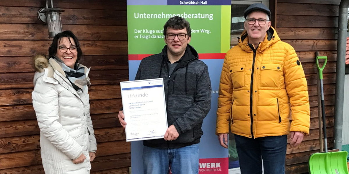 (von links) Christina Bauer, Markus Kretzschmar und Bernd Schöller. Drei Personen stehen nebeneinander und schauen in die Kamera, Mann in der Mitte hält eine gerahmte Urkunde in der Hand.