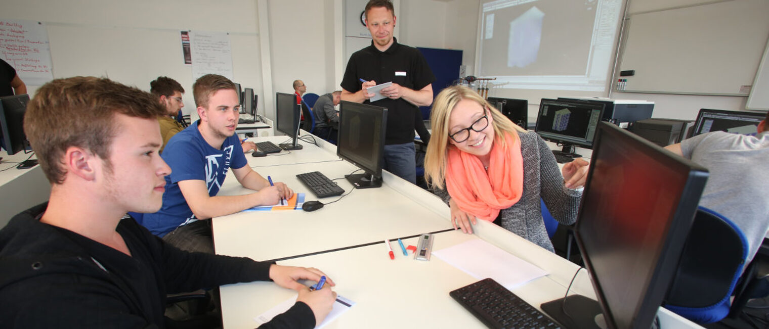 Schüler & Lehrer beim Unterricht in einem Raum mit Computern, im Vordergrund deutet eine Schülerin auf einen Monitor