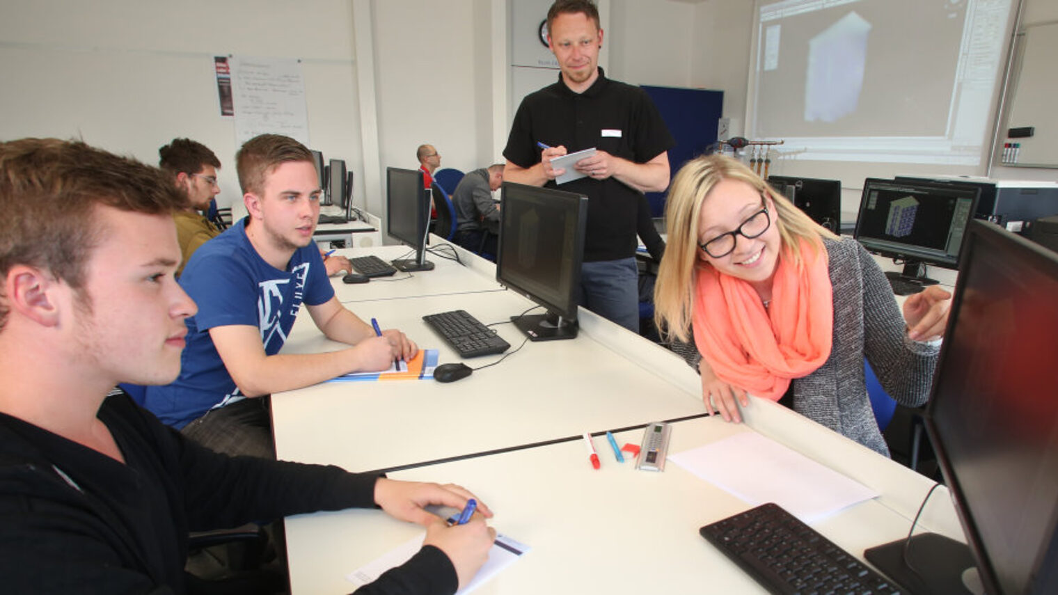 Schüler & Lehrer beim Unterricht in einem Raum mit Computern, im Vordergrund deutet eine Schülerin auf einen Monitor