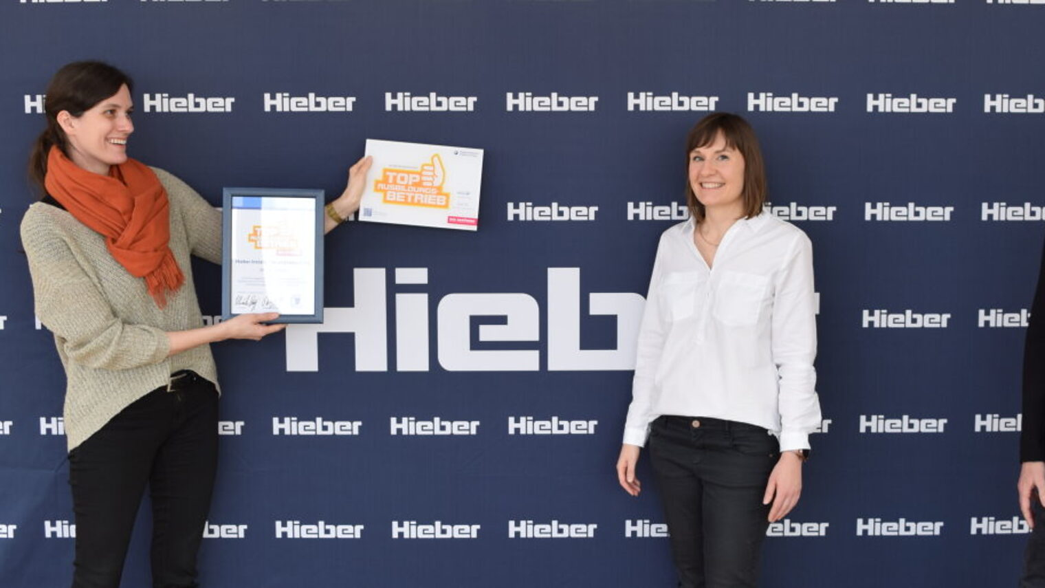 Frau überreicht einer Frau und einem Mann eine Urkunde und eine Plakette vor dunkelblauer Messewand mit Schriftzug Hieber