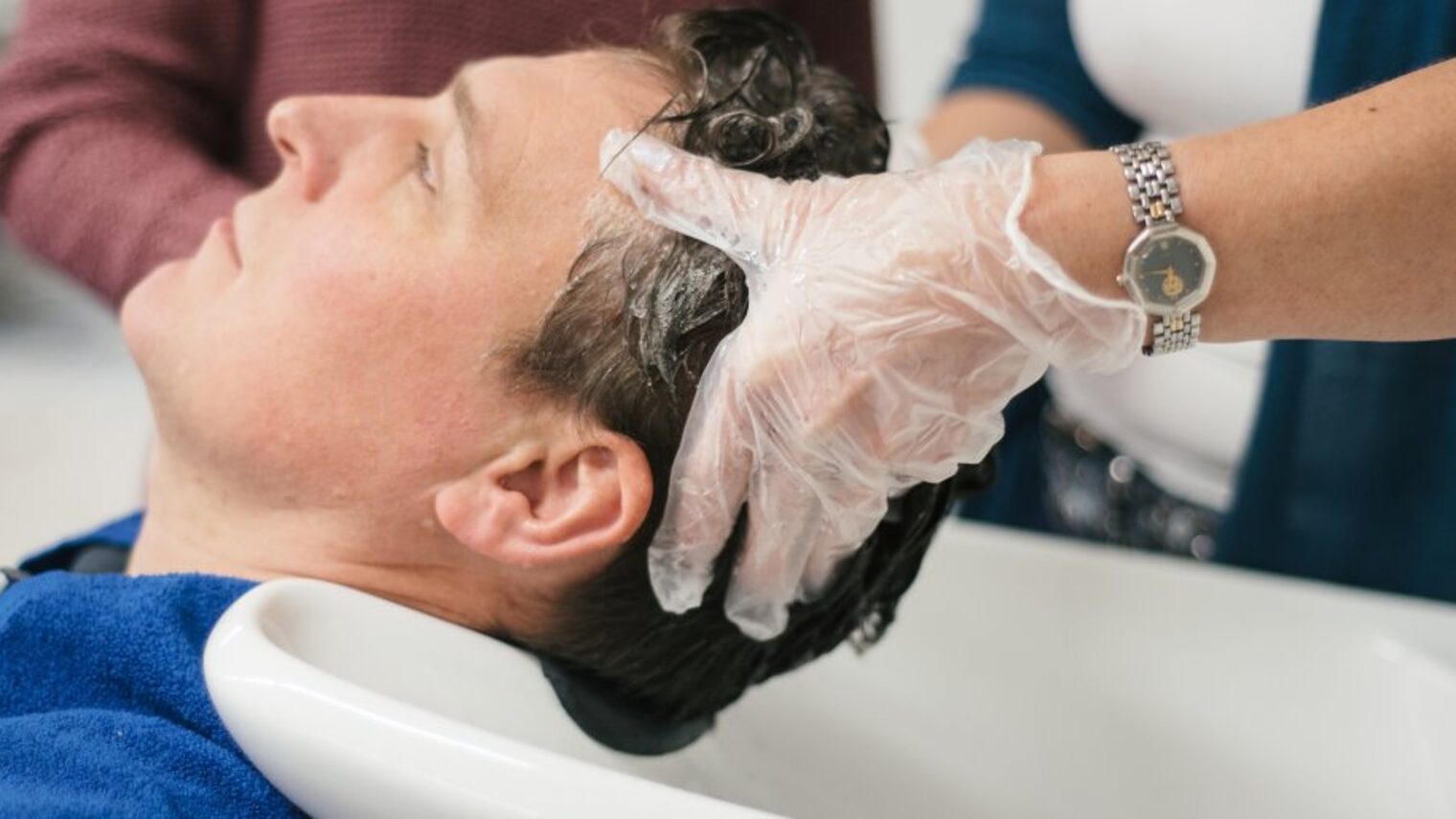 Einem Mann werden an einem Waschtisch die Haare gewaschen. Friseurin trägt dabei Handschuhe. 