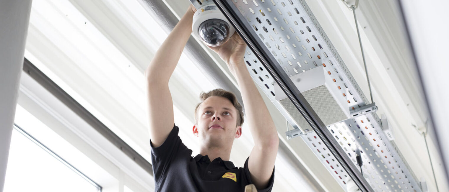 Bei den Elektro- und Metallberufen gab es 2021 mehr neue Lehrverträge. Junger Mann mit kurzen blonden Haaren und schwarzem T-Shirt bringt ein faustgroßes technisches Gerät an einer Deckenleiste an