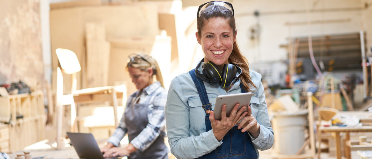 Web-Seminare für Handwerker - kostenfrei und wo Sie wollen. Lächelnde Handwerkerin in Tischler-Werkstatt mit Tablet-Computer in der Hand.