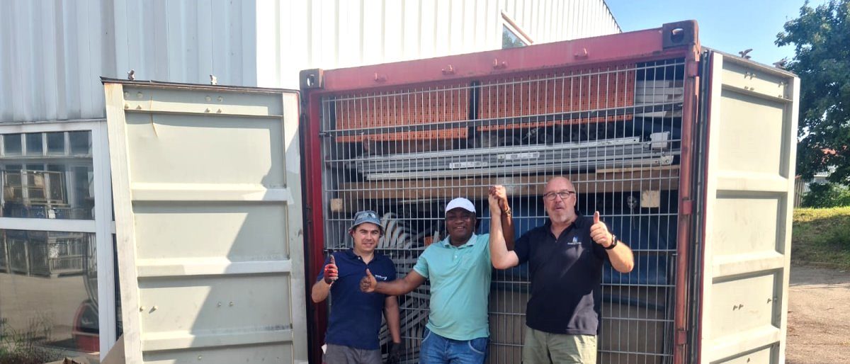 Karlheinz Mack (r.) und Mitarbeiter sammeln Spenden für Kamerun. Drei Männer stehen auf einem Betriebsgelände vor einem vollen Schiffscontainer mit offenen Toren und freuen sich.