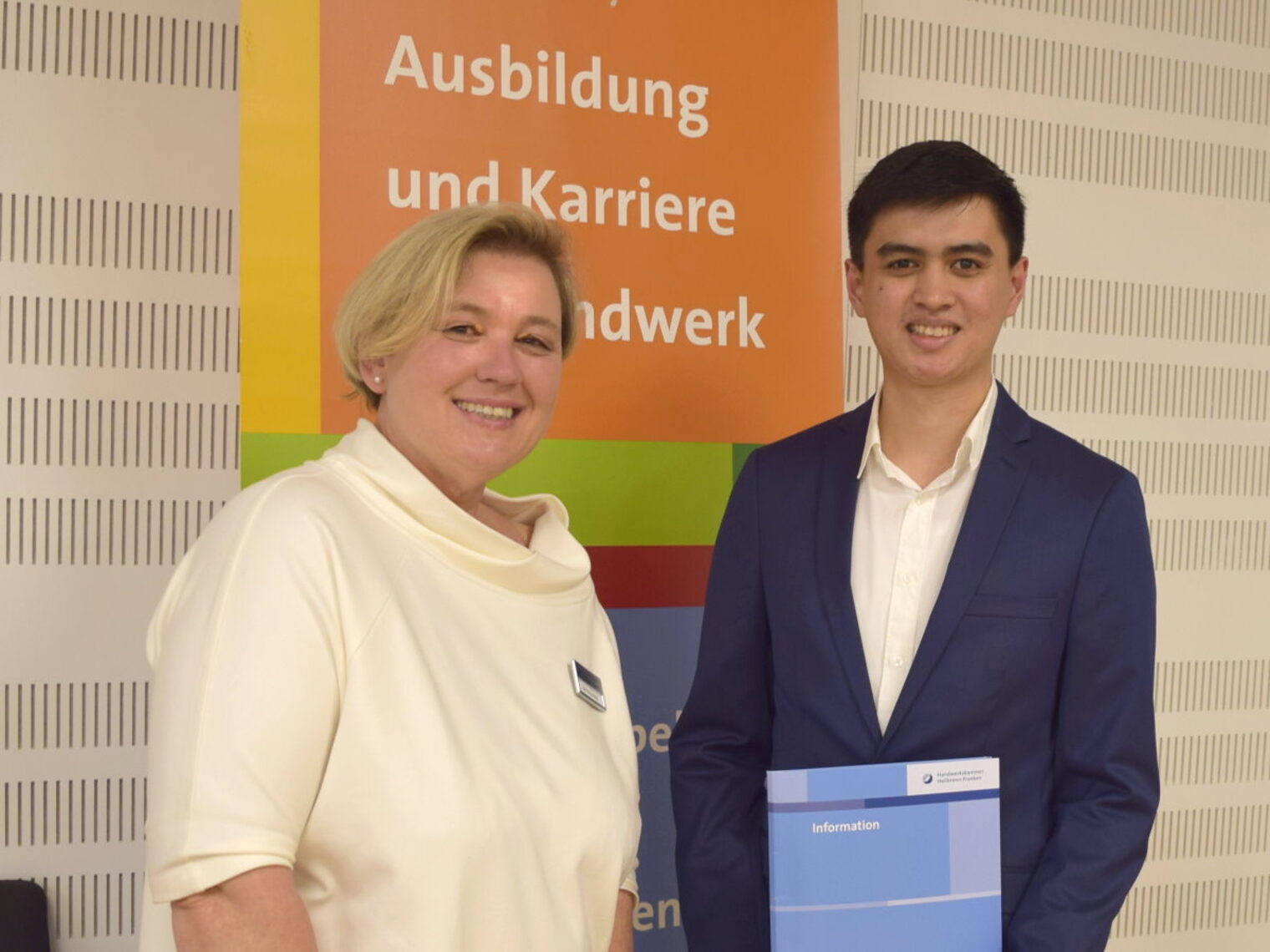 Eine Frau und ein junger Mann mit einer blauen Mappe stehen nebeneinander vor einem bunten Banner und lächeln in die Kamera.