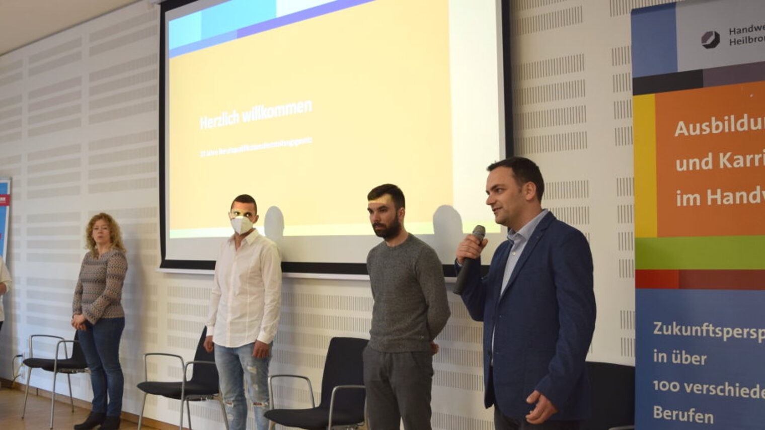 Zwei Frauen und drei Männer stehen nebeneinander in einem Raum vor einer Leinwand, Mann ganz rechts spricht in ein Mikrofon.