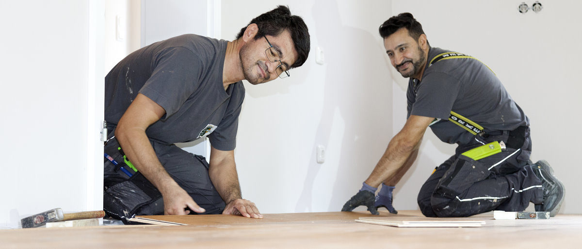 Im ersten Quartal des Jahres war die Stimmung im Handwerk noch gut. Zwei Männer in Arbeitskleidung verlegen knieend in einem Zimmer mit weißen Wänden einen Parkettboden.