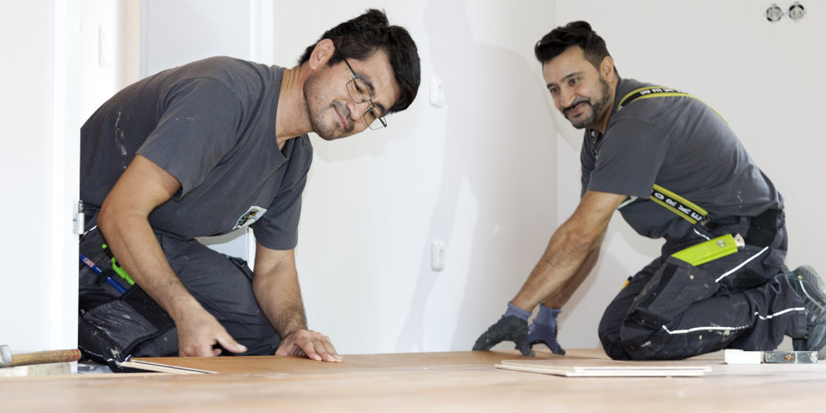 Zwei Männer in Arbeitskleidung verlegen knieend in einem Zimmer mit weißen Wänden einen Parkettboden.